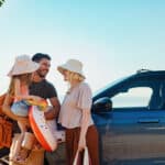 A cheerful family gathers near their car beside the sea, ready for a fun vacation. The sun shines brightly as they prepare with beach gear, symbolizing carefree summer days filled with joy.