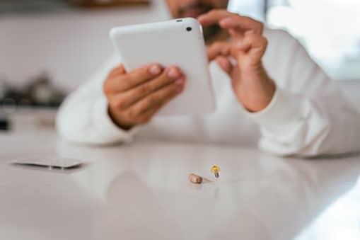Hearing aid sitting on a table; a man using his tablet in the background.